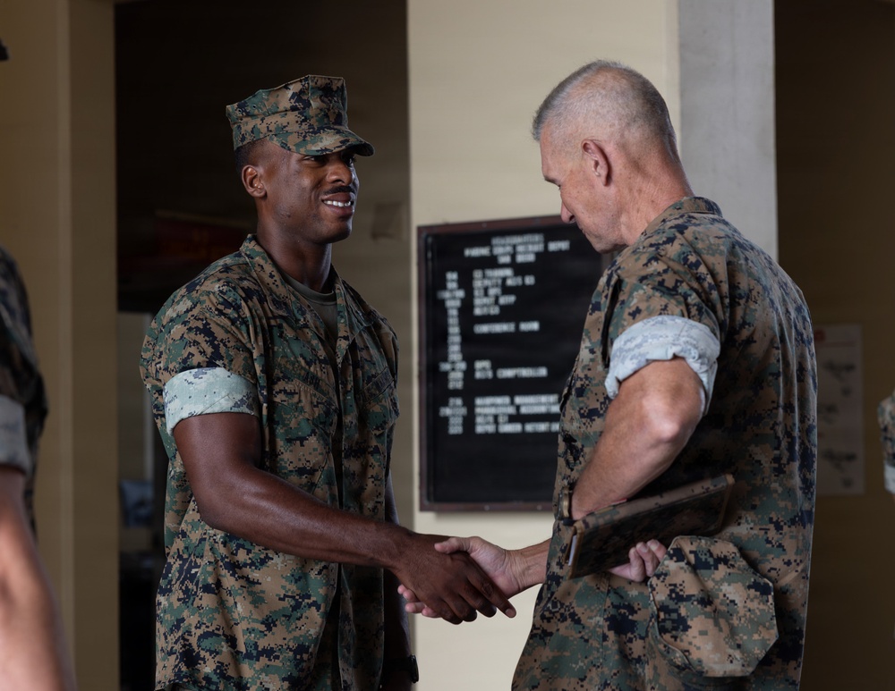 Two U.S. Marines in MARPAT woodland digital camouflage uniforms are shaking hands in what appears to be an indoor military facility. The younger Marine on the left is smiling, while the older Marine on the right is holding a clipboard or tablet. A bulletin board with a schedule or agenda is visible on the wall in the background.