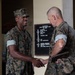 Two U.S. Marines in MARPAT woodland digital camouflage uniforms are shaking hands in what appears to be an indoor military facility. The younger Marine on the left is smiling, while the older Marine on the right is holding a clipboard or tablet. A bulletin board with a schedule or agenda is visible on the wall in the background.