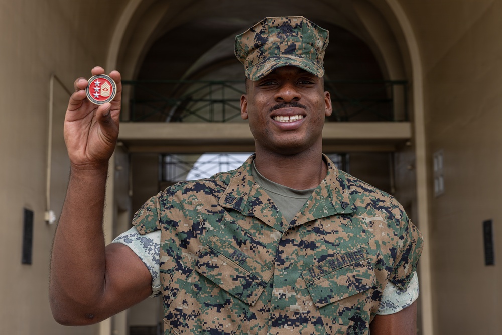 A U.S. Marine in MARPAT woodland camouflage uniform is holding up a military challenge coin for display. The Marine is smiling and standing in front of an arched building entrance. The challenge coin appears to feature a red design with stars, consistent with a senior enlisted or command-level coin.