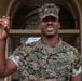 A U.S. Marine in MARPAT woodland camouflage uniform is holding up a military challenge coin for display. The Marine is smiling and standing in front of an arched building entrance. The challenge coin appears to feature a red design with stars, consistent with a senior enlisted or command-level coin.