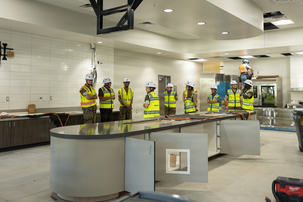 A group of approximately nine individuals wearing yellow high-visibility safety vests and white hard hats are gathered inside a large commercial kitchen or cafeteria facility under construction. Some individuals appear to be wearing military camouflage uniforms beneath their safety vests, suggesting a military inspection or oversight visit. In the background, an additional worker in an orange shirt is elevated on equipment, actively performing construction work.
