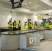 A group of approximately nine individuals wearing yellow high-visibility safety vests and white hard hats are gathered inside a large commercial kitchen or cafeteria facility under construction. Some individuals appear to be wearing military camouflage uniforms beneath their safety vests, suggesting a military inspection or oversight visit. In the background, an additional worker in an orange shirt is elevated on equipment, actively performing construction work.