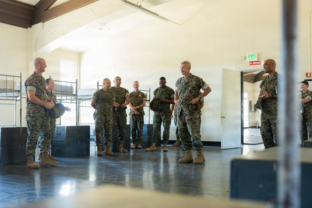 A group of approximately 8-10 military personnel in camouflage uniforms are gathered inside what appears to be a military barracks or training facility. They appear to be engaged in an informal briefing or conversation, with several individuals smiling and appearing relaxed. The setting includes bunk bed frames and storage equipment visible in the background.