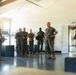 A group of approximately 8-10 military personnel in camouflage uniforms are gathered inside what appears to be a military barracks or training facility. They appear to be engaged in an informal briefing or conversation, with several individuals smiling and appearing relaxed. The setting includes bunk bed frames and storage equipment visible in the background.