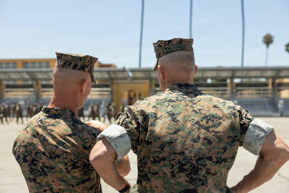 Two U.S. Marine Corps personnel are photographed from behind, observing what appears to be a formation or drill exercise on a parade deck. Both individuals are wearing MARPAT (Marine Pattern) digital camouflage uniforms and garrison covers (caps). The setting appears to be a military installation, likely a recruit depot or training facility, with bleachers and palm trees visible in the background.