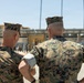 Two U.S. Marine Corps personnel are photographed from behind, observing what appears to be a formation or drill exercise on a parade deck. Both individuals are wearing MARPAT (Marine Pattern) digital camouflage uniforms and garrison covers (caps). The setting appears to be a military installation, likely a recruit depot or training facility, with bleachers and palm trees visible in the background.