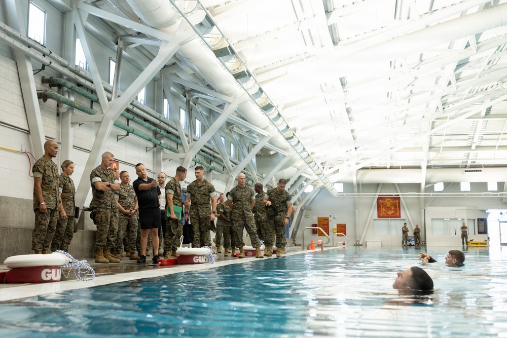A group of military personnel in camouflage uniforms, along with a few civilians, stand at the edge of an indoor swimming pool observing a water training exercise. Two swimmers are visible in the pool while the group on the poolside appears to be receiving a briefing or demonstration. The facility features a large industrial-style roof structure with white steel trusses and natural lighting.