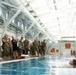 A group of military personnel in camouflage uniforms, along with a few civilians, stand at the edge of an indoor swimming pool observing a water training exercise. Two swimmers are visible in the pool while the group on the poolside appears to be receiving a briefing or demonstration. The facility features a large industrial-style roof structure with white steel trusses and natural lighting.