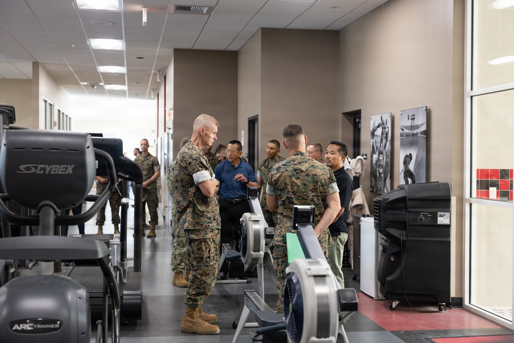 A group of U.S. Marines in woodland MARPAT camouflage uniforms are gathered in a modern military fitness facility, appearing to receive a briefing or tour of the equipment. Several civilians in casual business attire are also present among the group. The facility contains various cardio machines including rowing ergometers, an Arc Trainer, and Cybex equipment.