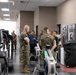 A group of U.S. Marines in woodland MARPAT camouflage uniforms are gathered in a modern military fitness facility, appearing to receive a briefing or tour of the equipment. Several civilians in casual business attire are also present among the group. The facility contains various cardio machines including rowing ergometers, an Arc Trainer, and Cybex equipment.