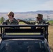 Two military personnel in US Navy Working Uniform (NWU) camouflage are positioned on the roof rack of a tactical vehicle in a rural, open landscape. One individual is gesturing toward the other, suggesting active communication or instruction. Two additional personnel are visible inside the vehicle through the windshield.