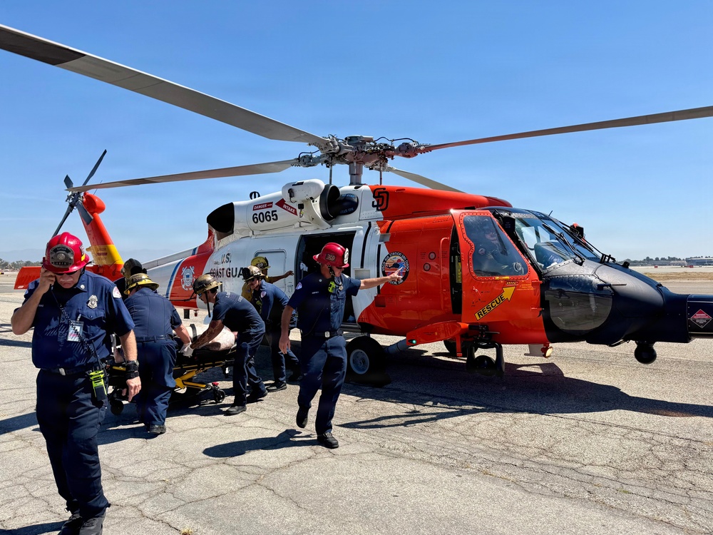 A U.S. Coast Guard helicopter (tail number 6065) is on the ground at what appears to be an airfield or tarmac. Emergency personnel in blue uniforms and red helmets are actively transferring a patient on a stretcher from the aircraft. The scene depicts a medical evacuation or medevac operation in progress under clear, sunny skies.