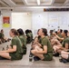 A female Marine drill instructor in camouflage utilities and campaign cover addresses a large group of female recruits seated on a concrete floor in what appears to be a military training facility. The recruits are wearing olive green PT (physical training) uniforms and have water bottles placed in front of them. A USMC Rank Structure chart and other military posters are visible on the walls in the background.