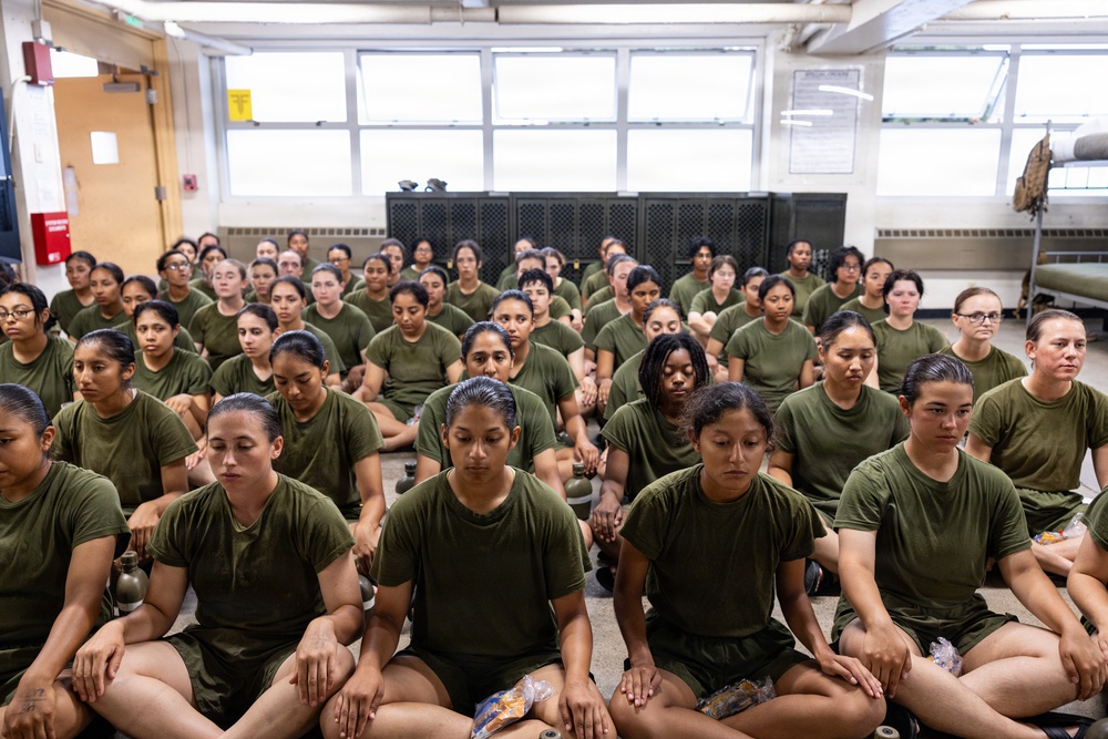 A large group of women dressed in identical olive green military-style t-shirts are seated on the floor in rows inside what appears to be a military training facility or barracks. Their expressions are serious and attentive, suggesting they are receiving instruction or waiting during a training session. The setting, uniforms, and disciplined posture are consistent with military recruit training.