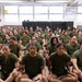 A large group of women dressed in identical olive green military-style t-shirts are seated on the floor in rows inside what appears to be a military training facility or barracks. Their expressions are serious and attentive, suggesting they are receiving instruction or waiting during a training session. The setting, uniforms, and disciplined posture are consistent with military recruit training.