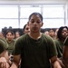 A group of individuals wearing olive green military-style t-shirts are seated cross-legged on a floor in an indoor setting. The foreground subject has wet hair and a serious expression, with a military canteen labeled 'SILVA B' and a packaged snack visible nearby. The setting and uniforms suggest a military training or recruit environment.
