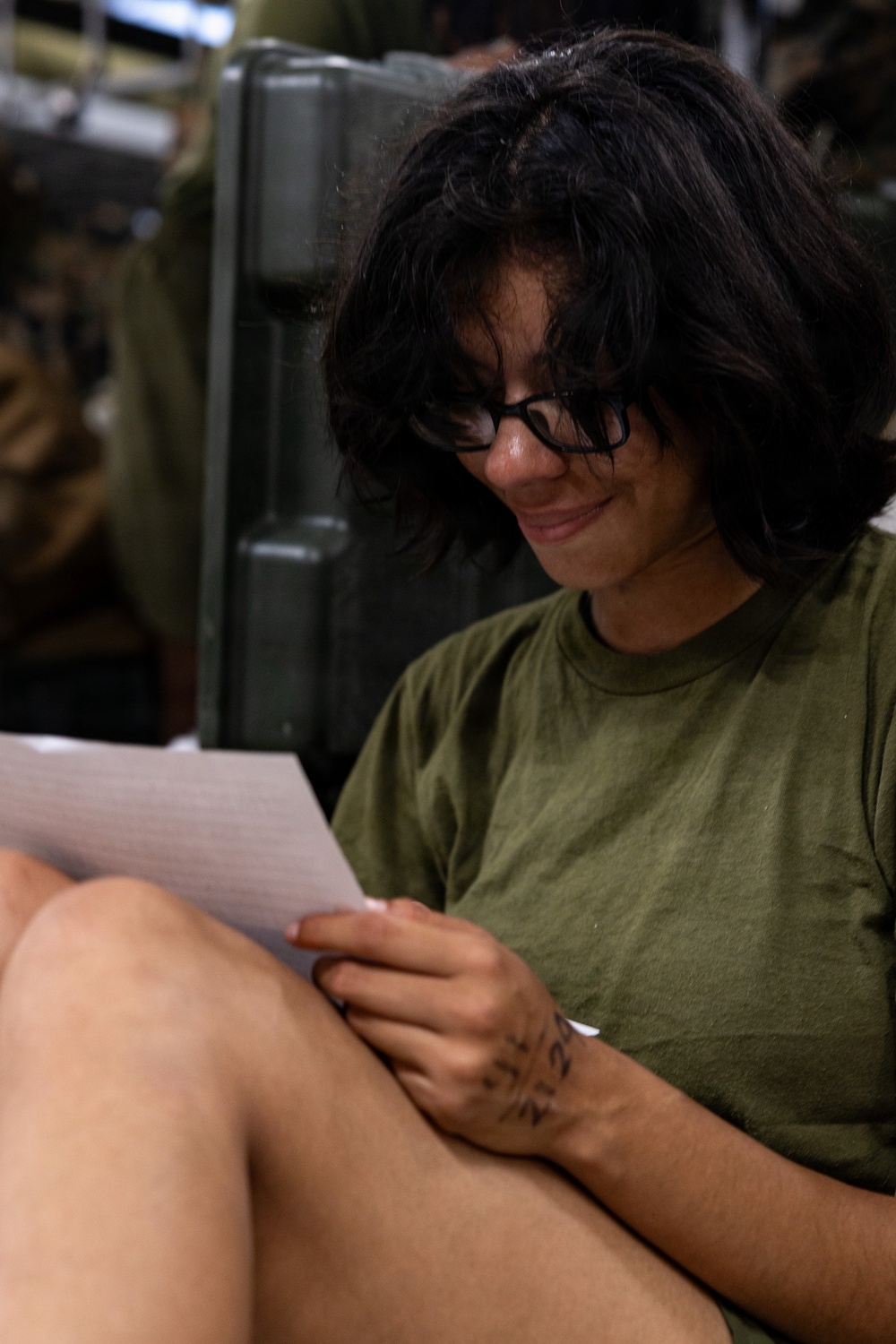 A person with dark hair and black-rimmed glasses is seated and reading a piece of paper or letter, smiling slightly. They are wearing an olive green t-shirt and have a visible tattoo on their hand. The background contains what appears to be military-style equipment or storage containers.
