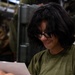 A person with dark hair and black-rimmed glasses is seated and reading a piece of paper or letter, smiling slightly. They are wearing an olive green t-shirt and have a visible tattoo on their hand. The background contains what appears to be military-style equipment or storage containers.