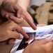 Two people are examining a printed photograph together, with both pointing at specific areas of the image. The photo appears to show an interior space with blue and red elements visible. The scene suggests a collaborative review or discussion of the printed material.