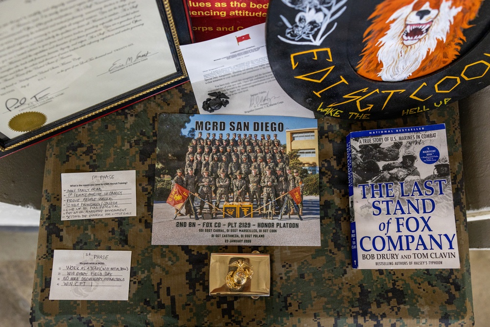 A flat-lay display of U.S. Marine Corps memorabilia and documents arranged on a camouflage surface. The items appear to belong to a Marine recruit or graduate from MCRD San Diego, including a platoon photo, personal goal notes, a military book, and various insignia. The collection documents a recruit's journey through Marine Corps boot camp circa January 2020.