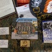 A flat-lay display of U.S. Marine Corps memorabilia and documents arranged on a camouflage surface. The items appear to belong to a Marine recruit or graduate from MCRD San Diego, including a platoon photo, personal goal notes, a military book, and various insignia. The collection documents a recruit's journey through Marine Corps boot camp circa January 2020.