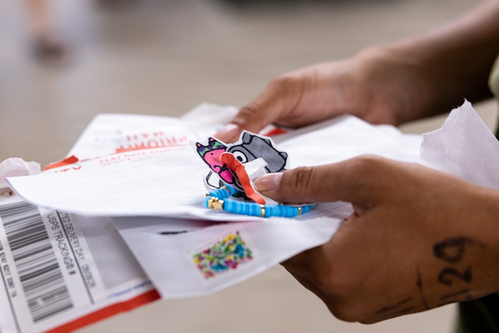 A person is holding a collection of mail items including what appears to be a USPS Priority Mail flat rate envelope and other correspondence. Among the mail items, there is a small cartoon character sticker/patch and a blue beaded bracelet visible. The image appears to capture someone sorting through recently received mail or packages.