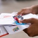 A person is holding a collection of mail items including what appears to be a USPS Priority Mail flat rate envelope and other correspondence. Among the mail items, there is a small cartoon character sticker/patch and a blue beaded bracelet visible. The image appears to capture someone sorting through recently received mail or packages.
