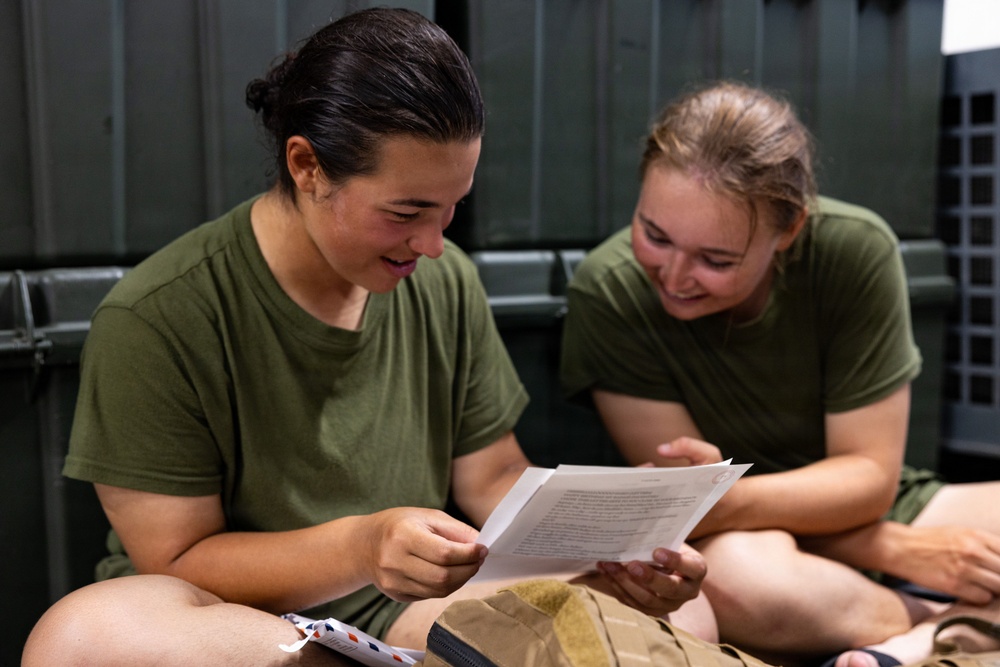 Two young women wearing matching olive green military-style t-shirts are seated on the floor, smiling while reading a letter or document together. The setting appears to be a military facility or barracks, suggested by the dark green metal lockers or equipment visible in the background. The mood is positive and candid, capturing a moment of shared enjoyment.