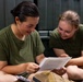 Two young women wearing matching olive green military-style t-shirts are seated on the floor, smiling while reading a letter or document together. The setting appears to be a military facility or barracks, suggested by the dark green metal lockers or equipment visible in the background. The mood is positive and candid, capturing a moment of shared enjoyment.