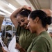 Two women wearing olive green military-style t-shirts are sharing a moment of joy while reading what appears to be a letter or document together. Both are smiling and laughing, suggesting the content is positive or amusing. The setting appears to be a military supply room or storage facility with shelving and boxes visible in the background.