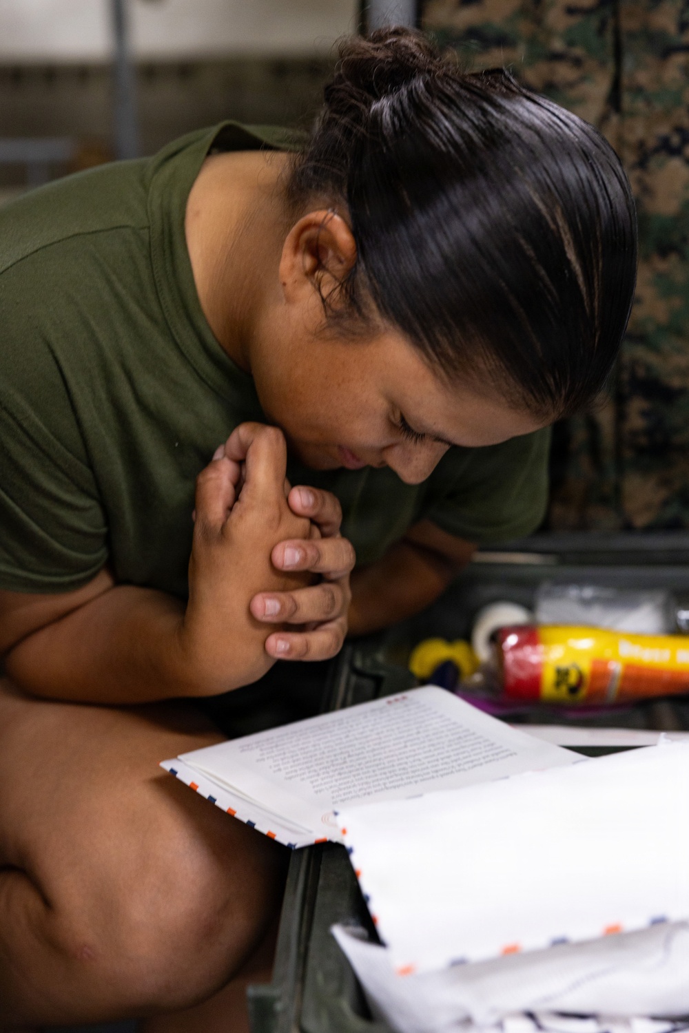 A person wearing an olive green military-style t-shirt is leaning forward with hands clasped, looking down at an open document or letter. Several papers with blue and orange border markings are stacked in front of them, suggesting military mail correspondence. A camouflage uniform is visible in the background, reinforcing a military setting.