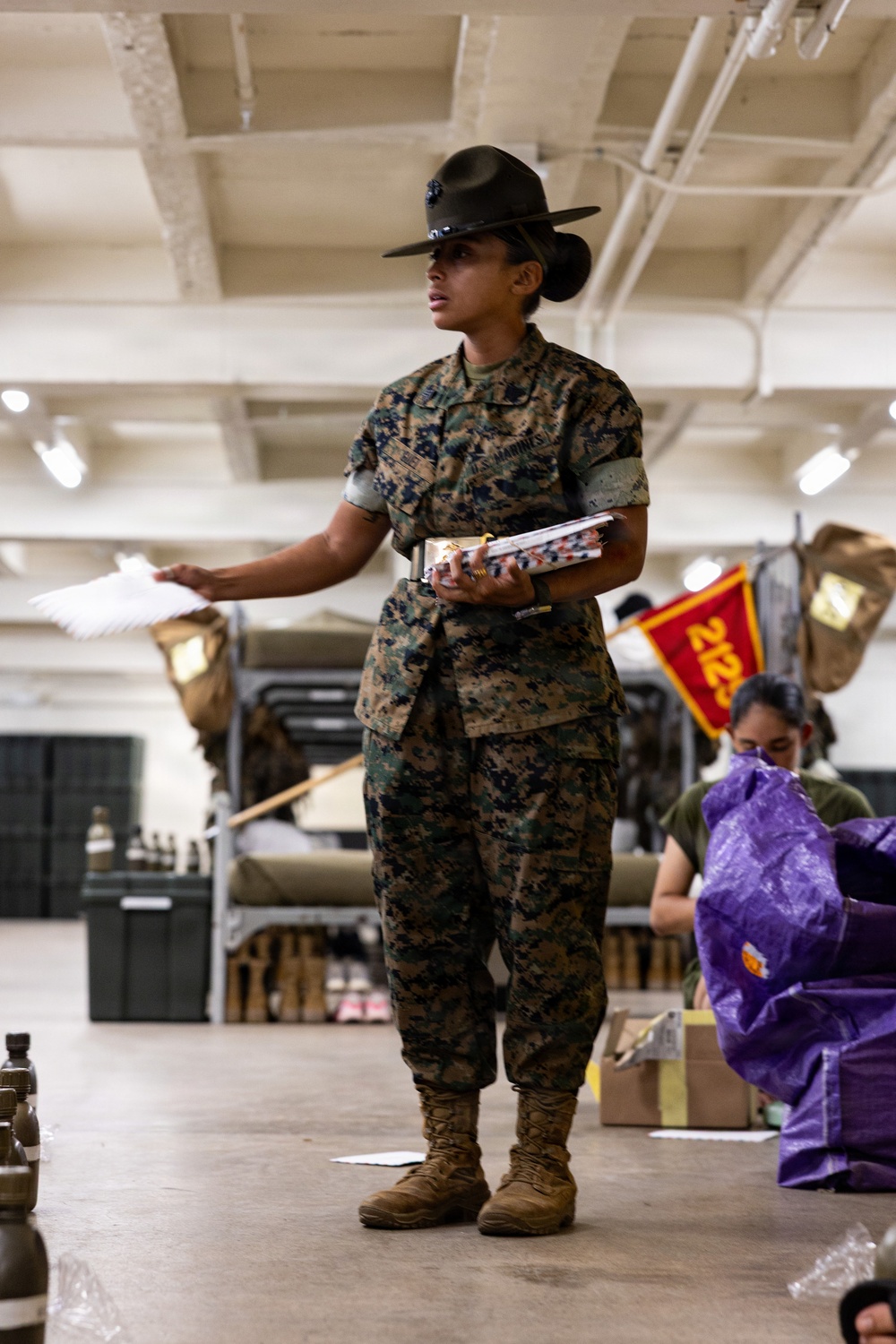 A U.S. Marine drill instructor wearing the distinctive campaign cover (drill sergeant hat) stands in what appears to be a warehouse or supply facility, holding documents and addressing personnel. She is dressed in MARPAT camouflage utilities with 'U.S. MARINES' tape visible on her uniform. In the background, another service member works with supplies near a red pennant bearing the number '2123'.
