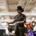 A U.S. Marine drill instructor wearing the distinctive campaign cover (drill sergeant hat) stands in what appears to be a warehouse or supply facility, holding documents and addressing personnel. She is dressed in MARPAT camouflage utilities with 'U.S. MARINES' tape visible on her uniform. In the background, another service member works with supplies near a red pennant bearing the number '2123'.