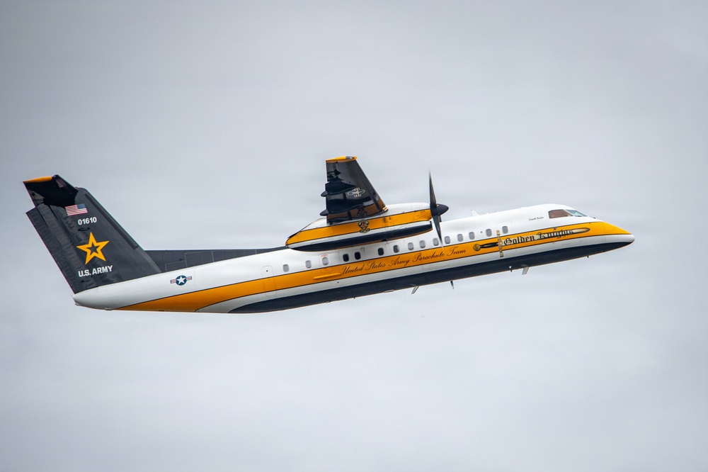 A U.S. Army turboprop aircraft belonging to the Golden Knights parachute team is captured in flight against an overcast sky. The aircraft is distinctively painted in white, black, and gold/yellow livery with U.S. Army markings. The tail number 01610 and 'U.S. Army' text are clearly visible on the tail section.