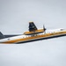 A U.S. Army turboprop aircraft belonging to the Golden Knights parachute team is captured in flight against an overcast sky. The aircraft is distinctively painted in white, black, and gold/yellow livery with U.S. Army markings. The tail number 01610 and 'U.S. Army' text are clearly visible on the tail section.