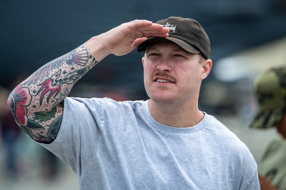 A young adult male is shielding his eyes from the sun with his right hand, as if looking into the distance. He is wearing a gray t-shirt and a dark baseball cap with the 'Stüssy' brand logo visible. His right arm features extensive traditional-style tattoos with floral and decorative motifs.