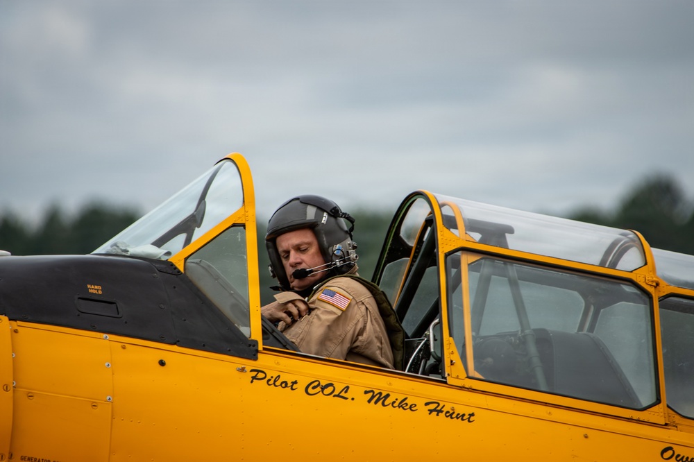 A pilot identified by fuselage text as 'Pilot COL. Mike Hunt' sits in the open cockpit of a vintage yellow aircraft, wearing a flight helmet with boom microphone and a tan flight suit with a US flag patch. The aircraft appears to be a classic military trainer, likely a North American T-6 Texan or similar WWII-era aircraft, based on its yellow paint scheme and cockpit design. The image was taken at what appears to be an airshow or airfield event under overcast skies.