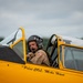 A pilot identified by fuselage text as 'Pilot COL. Mike Hunt' sits in the open cockpit of a vintage yellow aircraft, wearing a flight helmet with boom microphone and a tan flight suit with a US flag patch. The aircraft appears to be a classic military trainer, likely a North American T-6 Texan or similar WWII-era aircraft, based on its yellow paint scheme and cockpit design. The image was taken at what appears to be an airshow or airfield event under overcast skies.
