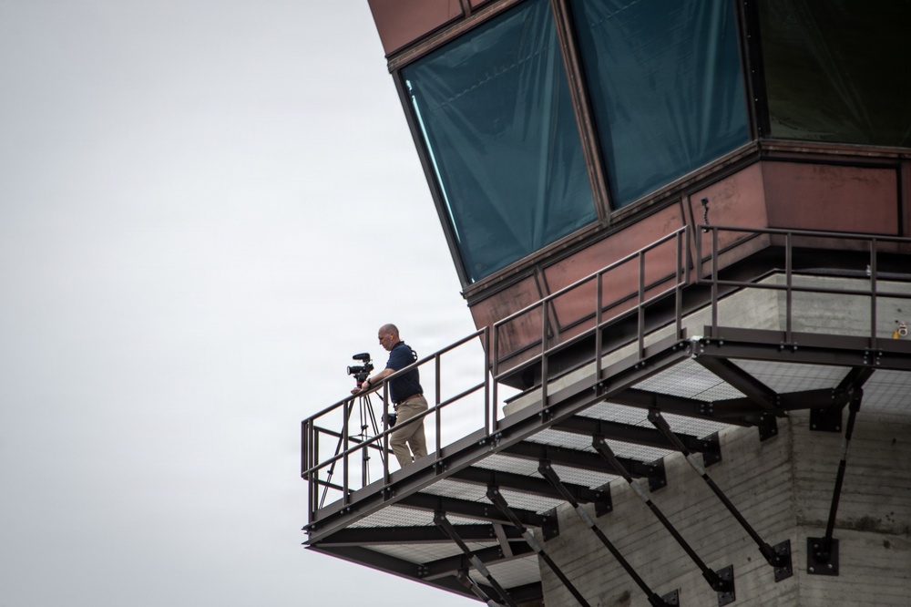 A bald man stands on an elevated metal walkway or observation platform attached to a large industrial or architectural structure. He is operating a camera mounted on a tripod, appearing to film or photograph from this elevated vantage point. The structure features large teal/green-covered windows or panels and steel railings.