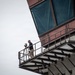 A bald man stands on an elevated metal walkway or observation platform attached to a large industrial or architectural structure. He is operating a camera mounted on a tripod, appearing to film or photograph from this elevated vantage point. The structure features large teal/green-covered windows or panels and steel railings.