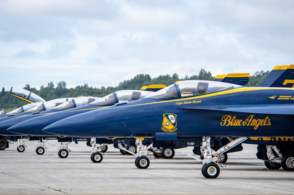 A line of U.S. Navy Blue Angels F/A-18 Super Hornet aircraft are parked on a tarmac in formation. The jets display their iconic navy blue and gold livery with 'Blue Angels' script and squadron insignia visible on the fuselages. The foreground aircraft is assigned to 'Capt Adam Bryan' as indicated by the name stenciled below the cockpit.