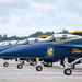 A line of U.S. Navy Blue Angels F/A-18 Super Hornet aircraft are parked on a tarmac in formation. The jets display their iconic navy blue and gold livery with 'Blue Angels' script and squadron insignia visible on the fuselages. The foreground aircraft is assigned to 'Capt Adam Bryan' as indicated by the name stenciled below the cockpit.