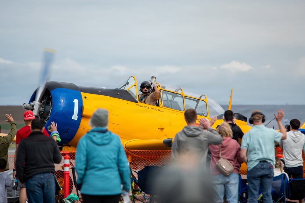 A yellow vintage military training aircraft, likely a North American T-6 Texan, is on the tarmac at an airshow. The pilot, identified by text on the fuselage as 'Pilot COL Mike Hunt,' is waving to a crowd of spectators gathered behind a safety fence. The crowd is enthusiastically waving back at the pilot in a celebratory exchange.