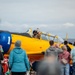 A yellow vintage military training aircraft, likely a North American T-6 Texan, is on the tarmac at an airshow. The pilot, identified by text on the fuselage as 'Pilot COL Mike Hunt,' is waving to a crowd of spectators gathered behind a safety fence. The crowd is enthusiastically waving back at the pilot in a celebratory exchange.