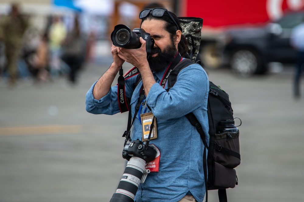 A bearded male photographer is actively shooting with a Canon DSLR camera raised to his eye. He is equipped with multiple cameras and professional gear, wearing a press/public affairs credential lanyard. The background is blurred, suggesting an outdoor public or media event.