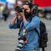 A bearded male photographer is actively shooting with a Canon DSLR camera raised to his eye. He is equipped with multiple cameras and professional gear, wearing a press/public affairs credential lanyard. The background is blurred, suggesting an outdoor public or media event.