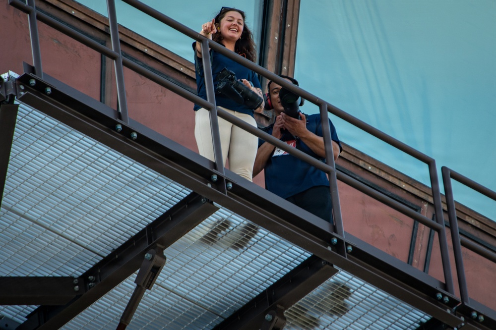 Two individuals are standing on a metal fire escape or elevated walkway, both holding DSLR cameras. The woman in the foreground is smiling and pointing upward, wearing a blue jacket and white pants with sunglasses on her head. The man behind her is looking down through his camera viewfinder.