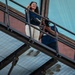 Two individuals are standing on a metal fire escape or elevated walkway, both holding DSLR cameras. The woman in the foreground is smiling and pointing upward, wearing a blue jacket and white pants with sunglasses on her head. The man behind her is looking down through his camera viewfinder.
