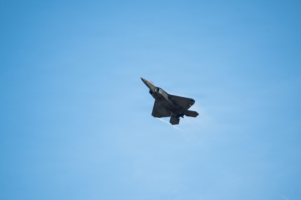 A stealth fighter jet, consistent with the design of an F-22 Raptor, is photographed in flight against a clear blue sky. The aircraft is captured in a banking maneuver, showing its underside and distinctive angular stealth geometry. A faint vapor trail or condensation effect is visible near the aircraft's body.