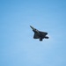 A stealth fighter jet, consistent with the design of an F-22 Raptor, is photographed in flight against a clear blue sky. The aircraft is captured in a banking maneuver, showing its underside and distinctive angular stealth geometry. A faint vapor trail or condensation effect is visible near the aircraft's body.